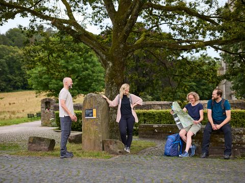 Vier Menschen machen Pause am Etappenstein in Odenthal-Altenberg. Zwei stehen direkt neben dem Stein und zwei sitzen und schauen in eine Wanderkarte. Im Hintergrund ist ein Stück vom Altenberger Dom zu erkennen. 