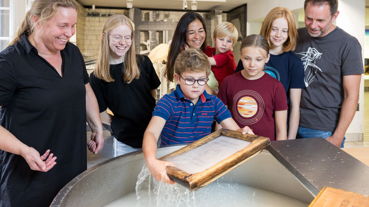 Einige Erwachsene, Kinder und Jugendliche stehen um ein Rundes Becken in dem ein Junge Papier schöpft.