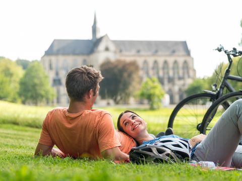 Ein Mann liegt auf einer Wiese neben einem Fahrrad. Eine Frau liegt neben ihm und hat den Kopf auf seinem Bauch abgelegt. Neben den beiden liegen zwei Helme auf dem Gras und im Hintergrund ist unscharf der Altenberger Dom zu sehen. 